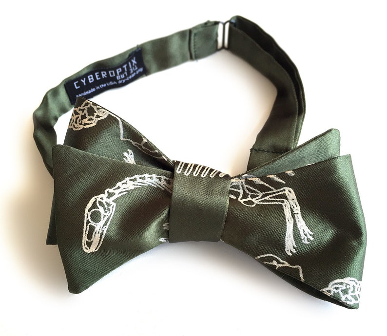 May include: A green bow tie with a white skeleton print. The bow tie is made of satin and has a  "CYBEROPTIX" label.