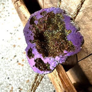 May include: A purple and gold bolo tie featuring a geode slice pendant. The pendant has a rough, natural edge and a sparkling, crystalline interior. The cord is a braided gold color, adding a touch of elegance to this unique accessory.