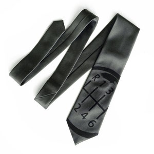 May include: A dark grey silk tie with a black printed gear shift pattern. The gear shift pattern includes the numbers 1, 2, 3, 4, 5, and 6, as well as the letter R.