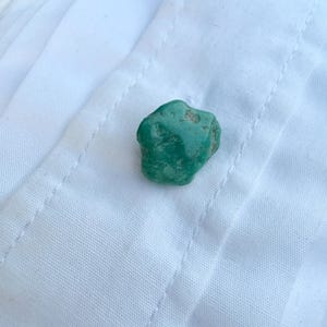 May include: A small, irregularly shaped, teal-colored stone rests on a white fabric surface. The stone has a rough texture and varying shades of green. The fabric has a subtle pattern and visible stitching.