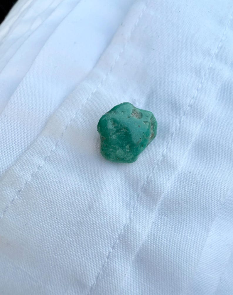 May include: A small, irregularly shaped, teal-colored stone rests on a white fabric surface. The stone has a rough texture and varying shades of green. The fabric has a subtle pattern and visible stitching.