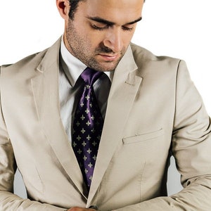 May include: A man wearing a beige suit jacket with a purple tie featuring a repeating pattern of gold fleur-de-lis. He is looking down at his wrist.