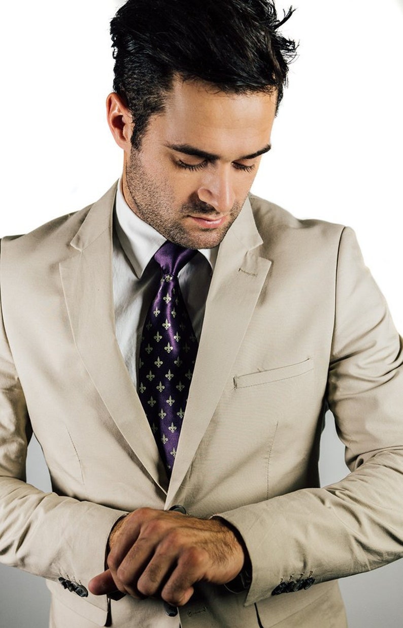 May include: A man wearing a beige suit jacket with a purple tie featuring a repeating pattern of gold fleur-de-lis. He is looking down at his wrist.