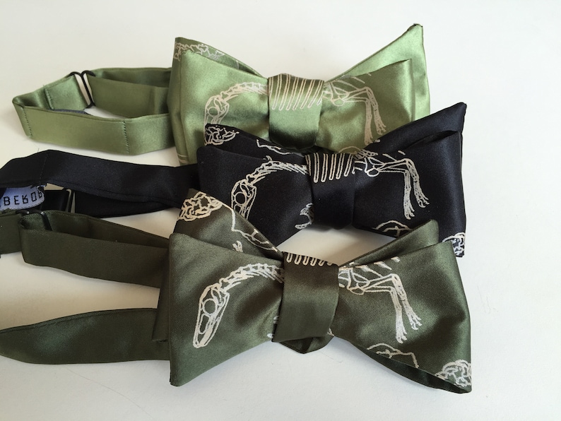 May include: Three bow ties with a dinosaur skeleton print. The bow ties are in shades of green and black.