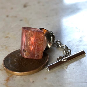 May include: A pink and orange crystal on a silver tie tack with a chain. The crystal is sitting on top of a US penny.
