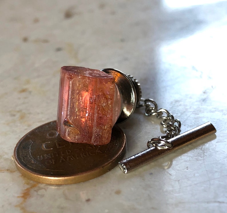 May include: A pink and orange crystal on a silver tie tack with a chain. The crystal is sitting on top of a US penny.