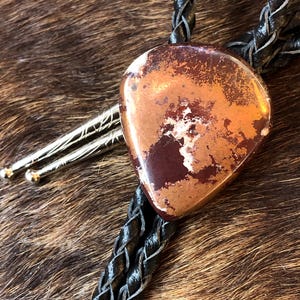 Peut inclure: Un bolo tie avec un cordon en cuir marron et un pendentif en forme de cœur argenté et cuivré. Le pendentif présente un motif marbré de brun, de cuivre et de blanc.