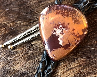 May include: A bolo tie with a brown leather cord and a silver and copper heart-shaped pendant. The pendant has a marbled pattern of brown, copper, and white.