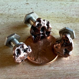 May include: Three copper-colored, textured stud earrings with silver-toned posts and hexagonal backs. The earrings are next to a penny coin. The earrings have a unique, organic shape, and are likely handmade.