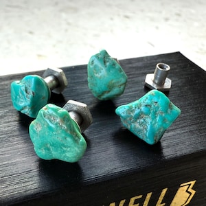May include: Four turquoise stone cabinet knobs with silver hardware are arranged on a black surface. The knobs have a natural, irregular shape and a vibrant blue-green color. The hardware is a silver-colored metal.