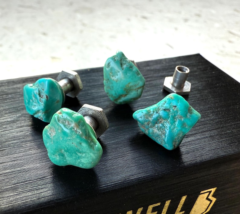 May include: Four turquoise stone cabinet knobs with silver hardware are arranged on a black surface. The knobs have a natural, irregular shape and a vibrant blue-green color. The hardware is a silver-colored metal.