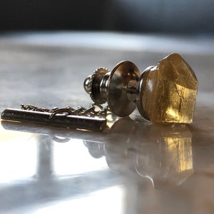 May include: A silver tie tack with a clear, faceted, yellow crystal. The tie tack has a chain attached to it.