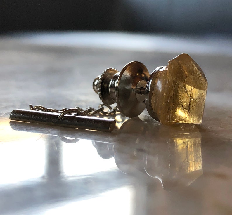 May include: A silver tie tack with a clear, faceted, yellow crystal. The tie tack has a chain attached to it.