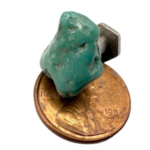 May include: A turquoise gemstone cabochon with a silver-colored metal post, resting on a copper penny. The turquoise is a vibrant teal color with natural variations. The penny is dated 1994.
