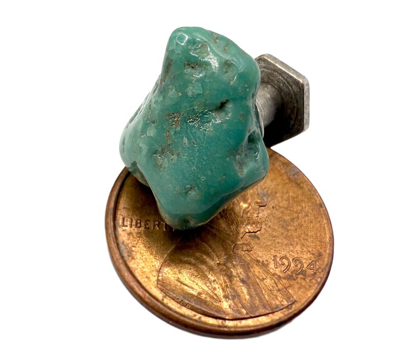 May include: A turquoise gemstone cabochon with a silver-colored metal post, resting on a copper penny. The turquoise is a vibrant teal color with natural variations. The penny is dated 1994.