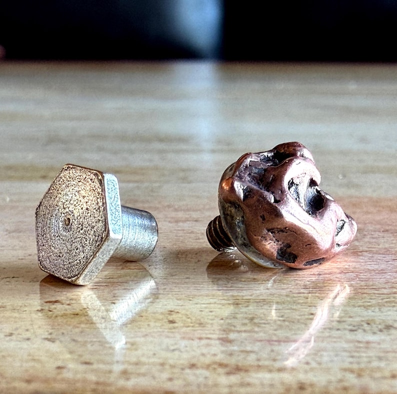 May include: Two metal knobs, one silver and hexagonal, the other a textured copper-colored shape. The silver knob has a textured surface and a cylindrical post. The copper knob has a screw base. Both are likely furniture hardware.
