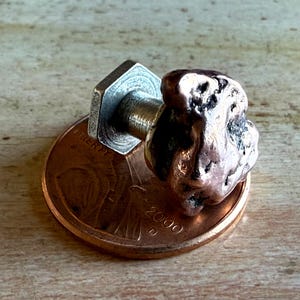 May include: A handcrafted copper and silver metal button with a unique, organic shape. The button is attached to a silver metal backing and sits on top of a copper penny. The penny has the year 2000 printed on it.