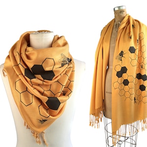 May include: A yellow scarf with a black honeycomb pattern and bee silhouettes. The scarf has fringe on the ends.