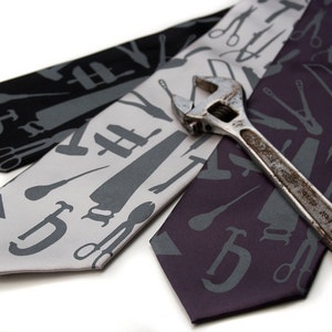 May include: Three silk ties with a pattern of tools in grey on a white, grey, and purple background. A silver wrench is lying on top of the ties.