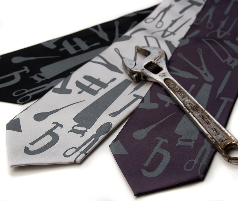May include: Three silk ties with a pattern of tools in grey on a white, grey, and purple background. A silver wrench is lying on top of the ties.