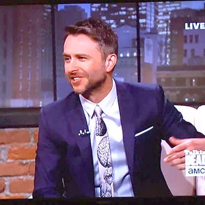 May include: A man wearing a dark blue suit jacket with a white shirt and a patterned tie. He is sitting in a chair with a white cushion. The background is a brick wall and a window with a city view. The text "Talking Dead" and "AMC" are visible on the chair.