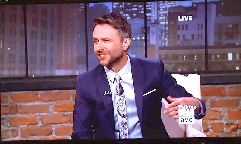 May include: A man wearing a dark blue suit jacket with a white shirt and a patterned tie. He is sitting in a chair with a white cushion. The background is a brick wall and a window with a city view. The text "Talking Dead" and "AMC" are visible on the chair.