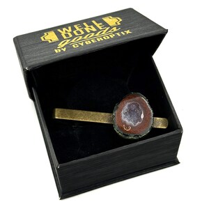 May include: A brass-toned metal tie bar featuring a raw, circular geode slice with a dark brown outer edge and a sparkling, light purple crystalline center. The tie bar is presented inside a black box with the gold text WELL DONE goods by CYBEROPTIX.
