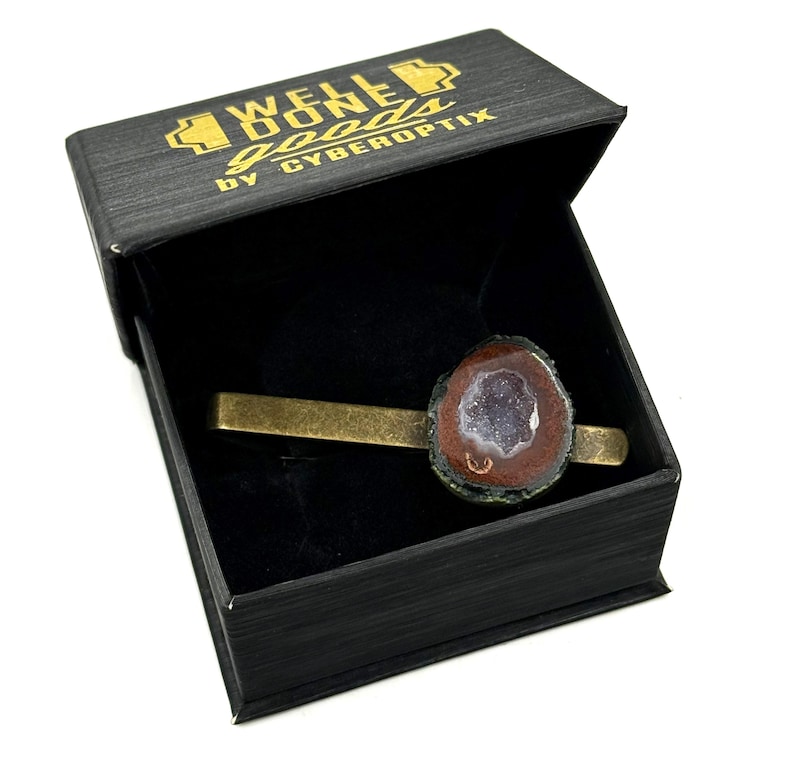 May include: A brass-toned metal tie bar featuring a raw, circular geode slice with a dark brown outer edge and a sparkling, light purple crystalline center. The tie bar is presented inside a black box with the gold text WELL DONE goods by CYBEROPTIX.