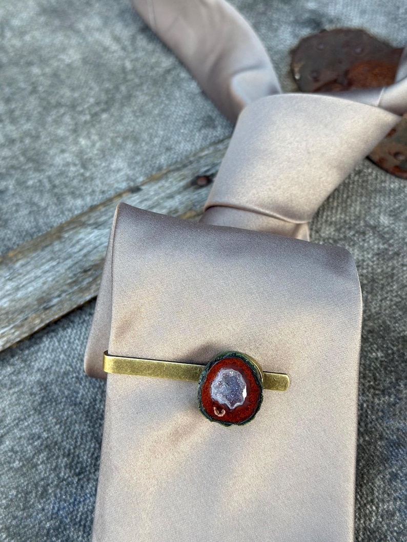 May include: A brass-toned metal tie clip featuring a polished, oval-shaped stone accent. The stone has a deep reddish-brown outer edge and a textured, crystalline center with light purple and white mineral deposits, secured to a smooth satin tie.