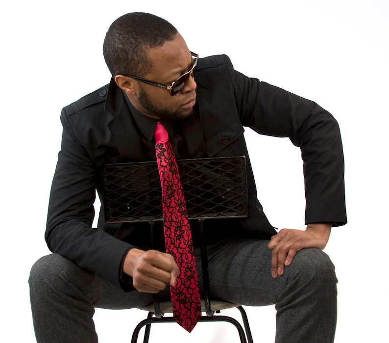 May include: A man wearing a black blazer, a black shirt, a red tie with black floral pattern, and gray pants. He is sitting on a black metal chair and wearing sunglasses.