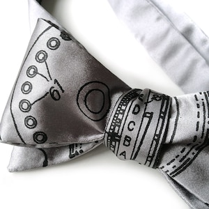 May include: A silver bow tie with a black and white geometric pattern. The pattern includes circles, lines, and letters.