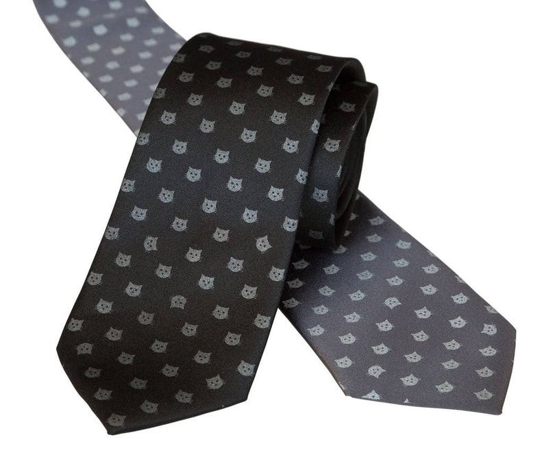 Puede incluir: Dos corbatas con un patr&oacute;n repetido de caras de gatos grises sobre un fondo gris oscuro. Una corbata est&aacute; enrollada y la otra est&aacute; extendida.