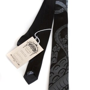 Puede incluir: Una corbata de seda negra con un dise&ntilde;o de pulpo gris. La corbata tiene una etiqueta que dice "The Verbosity Tie Lab" y "Hand Made in the USA".