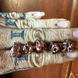 May include: Five copper-colored stud earrings with a textured, organic nugget design. The earrings have a natural, irregular shape and a metallic sheen. The earrings are displayed on a hand.