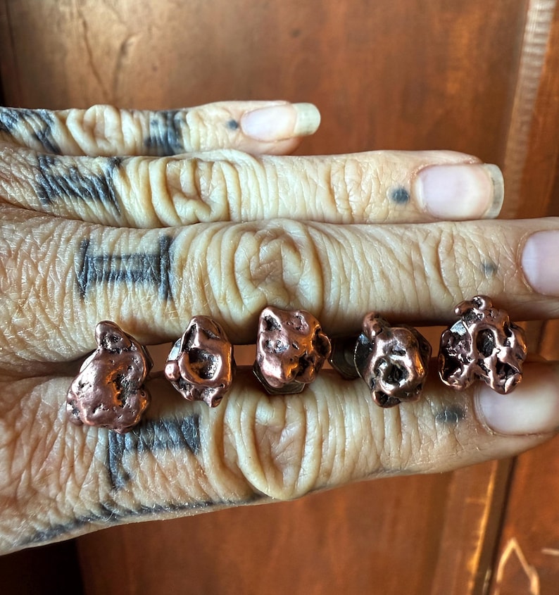 May include: Five copper-colored stud earrings with a textured, organic nugget design. The earrings have a natural, irregular shape and a metallic sheen. The earrings are displayed on a hand.