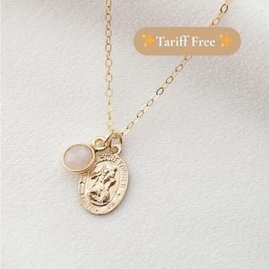 May include: Gold-colored necklace with a small, round, light-colored stone and a Saint Christopher pendant. The pendant has an oval shape with an image of Saint Christopher and the words "CHRISTOPHER PROTECT US". The necklace is on a white background.