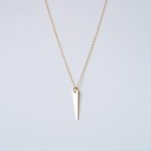 Puede incluir: Un delicado collar dorado con un colgante en forma de triángulo delgado y alargado. El colgante cuelga de una fina cadena sobre un fondo blanco. El collar es una pieza de joyería sencilla y elegante.