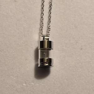 May include: A silver-colored necklace with a clear, cylindrical pendant. The pendant has silver-colored metal caps on the top and bottom, and a clear glass center. The chain is made of small, linked silver-colored metal pieces.