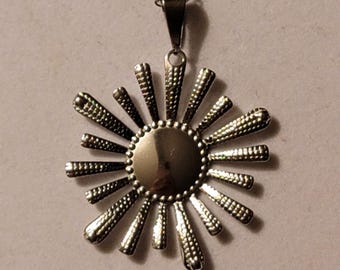 Puede incluir: Un collar de plata con un colgante de sol. El colgante tiene un centro circular y puntos radiantes.