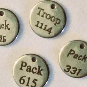 May include: Four round metal tags with holes for hanging. The tags are silver and have black text. The text on the tags reads "Pack 615", "Pack 337", "Troop 1114", and "Pack 25".