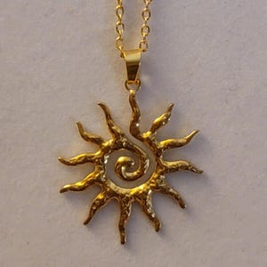 May include: A gold necklace with a sun pendant. The pendant is a stylized sun with a spiral in the center. The sun has a textured surface.