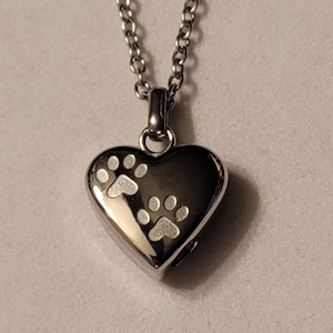May include: A silver heart-shaped pendant necklace with two paw prints engraved on the front. The chain is silver and has a delicate link design.