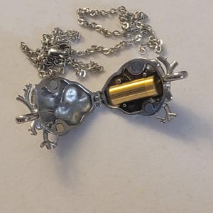 Op de afbeelding: Een zilveren ketting met een scharnierende hanger in de vorm van een hert. De hanger is open, waardoor een kleine gouden cilinder zichtbaar is.
