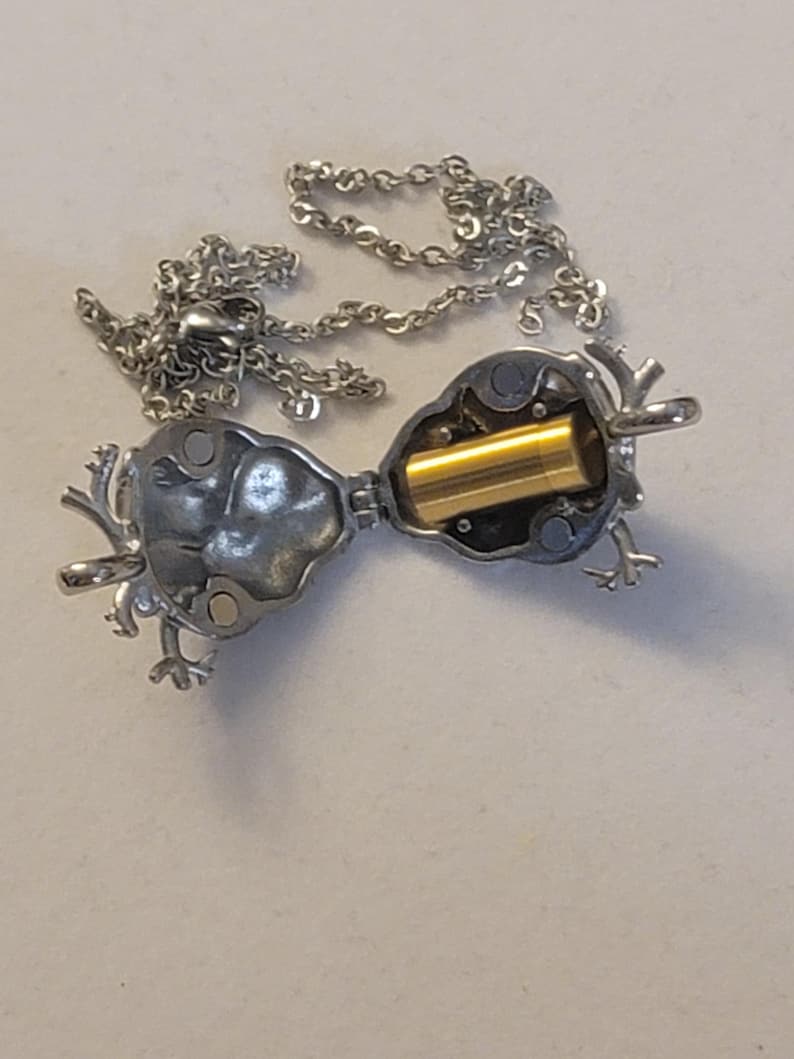 Op de afbeelding: Een zilveren ketting met een scharnierende hanger in de vorm van een hert. De hanger is open, waardoor een kleine gouden cilinder zichtbaar is.