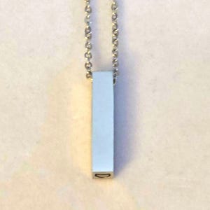 May include: A silver-colored pendant necklace featuring a rectangular bar pendant suspended from a delicate ball chain. The pendant has a smooth, reflective surface and a small, oval-shaped hole at the bottom. The necklace is simple and minimalist.
