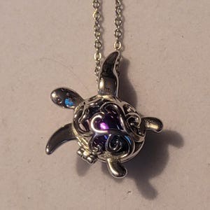 May include: A silver turtle pendant necklace. The turtle pendant features a detailed, openwork design with a purple iridescent interior. The necklace chain is also silver and delicate.