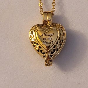 May include: Gold heart-shaped locket necklace with a delicate chain. The locket has an intricate design and the inscription "Always in my Heart".