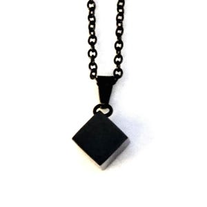 May include: A black necklace with a square pendant. The pendant is a solid, dark square shape, suspended from a matching black chain. The chain is made of small, linked circles. The background is plain white.