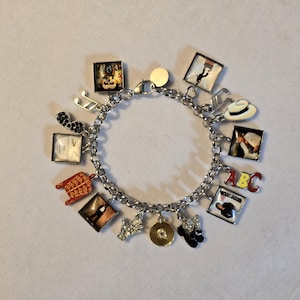 May include: A silver charm bracelet with various charms, including a record, a hat, and a square charm with the letters "ABC". Other charms include musical notes, a glove, and a silhouette of a person.