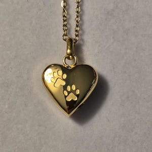 May include: Gold heart-shaped locket necklace with a chain. The locket has two paw prints engraved on the front.